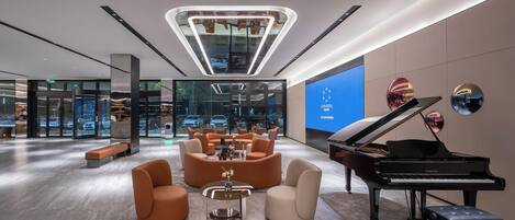 A modern lobby with a grand piano, comfortable seating, a central light fixture, and a large digital screen displaying the brand name.