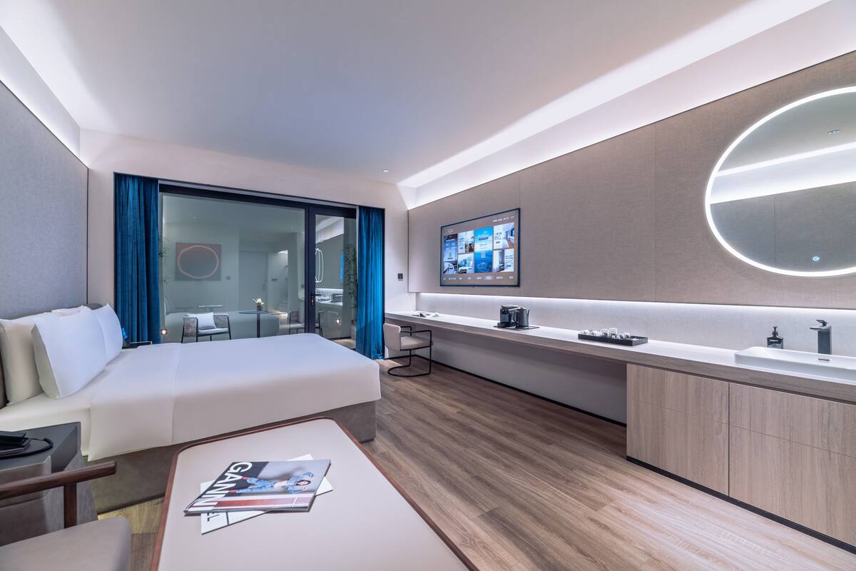 Une chambre d’hôtel moderne équipée d’un lit, d’un bureau, d’une table basse, d’une télévision et d’une salle de bain visible à travers une porte en verre.