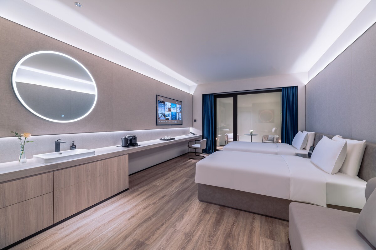 Een moderne hotelkamer met een groot bed, een ronde spiegel, een ingebouwde televisie, een wastafel met een wasbak en een groot raam met gordijnen.