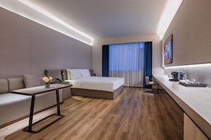 Een moderne hotelkamer met een bed, een bank, een klein tafeltje, een kitchenette en een groot raam met gordijnen.