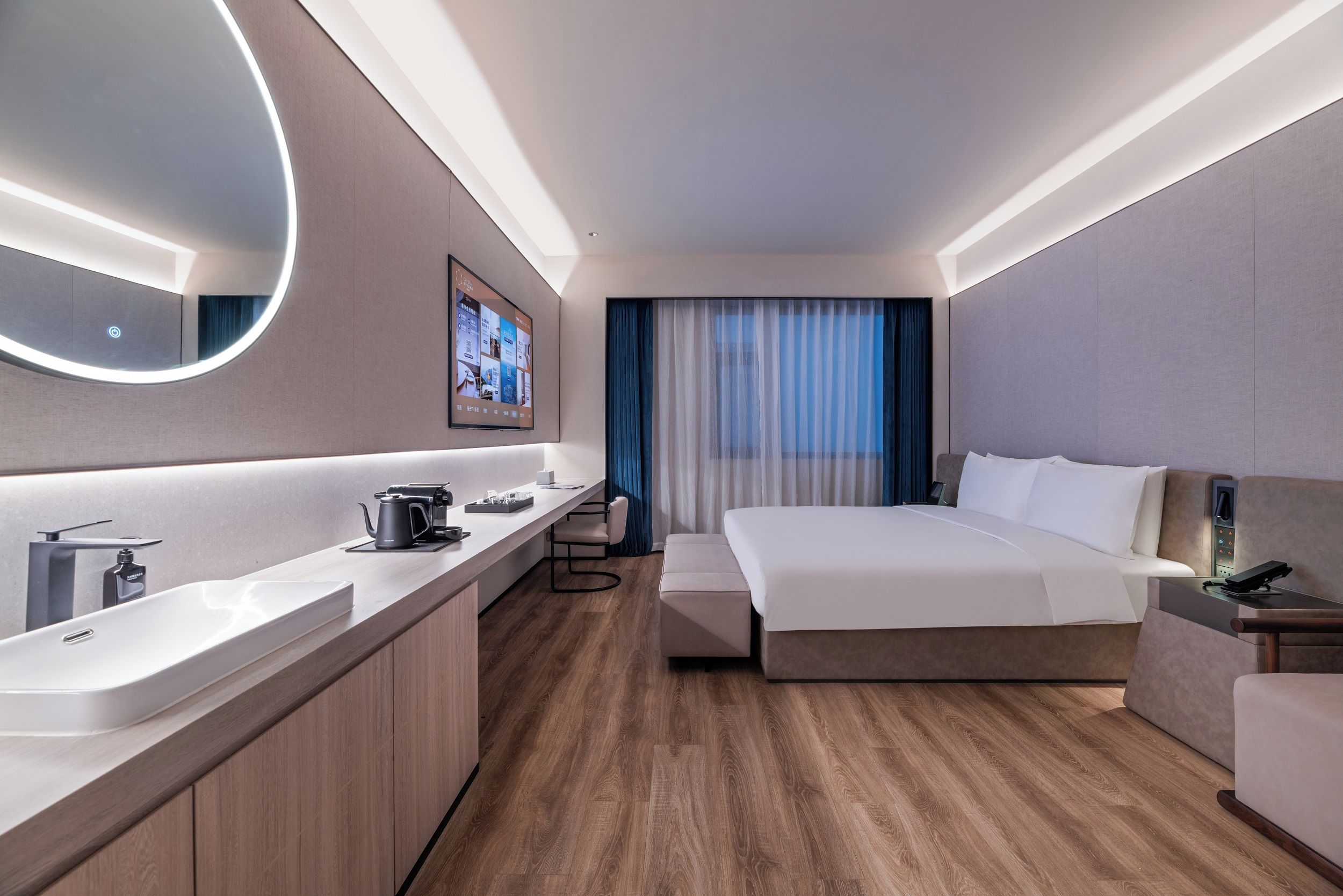 A modern hotel room with a large bed, a bedside table, a TV mounted on the wall, a bathroom with a sink and mirror, and a wooden floor.