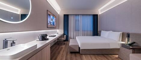 A modern hotel room with a large bed, a bedside table, a TV mounted on the wall, a bathroom with a sink and mirror, and a wooden floor.