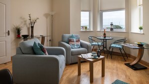 A living room with a grey sofa, a matching armchair, a small round table with a bottle and glasses, and a view of the sea through the windows. - Puffin View - One Bedroom Cottage, Sleeps 2 (Llanfairfechan)