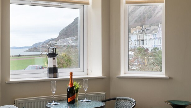 A room with a table by a window overlooking a coastal view, a bottle of wine, and two glasses. - Puffin View - One Bedroom Cottage, Sleeps 2 (Llanfairfechan)