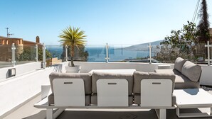 Una terraza en la azotea con un sofá blanco, una planta en maceta y una vista despejada del mar y colinas lejanas.