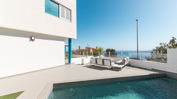Un área de piscina exterior moderna con vistas despejadas al mar, un edificio blanco con ventana azul y una farola.