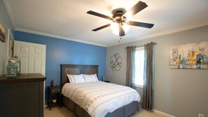 Un dormitorio con cama, mesita de noche, cómoda, ventilador de techo, ventana con cortinas y una obra de arte en la pared.