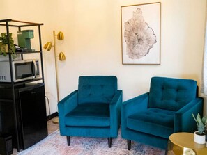 Une pièce comprenant deux fauteuils turquoise, un meuble noir pour appareils électroniques avec un micro-ondes, un lampadaire doré et une œuvre d’art abstraite encadrée accrochée au mur.