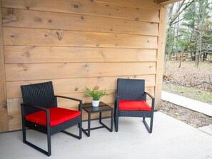 Deux fauteuils en osier noir avec des coussins rouges et une petite table sur laquelle est posée une plante en pot, le tout placé devant un mur en bois sur un sol en béton.