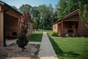 Une rangée de cabanes en bois, disposées de part et d’autre d’un chemin central, et entourées de verdure et d’arbres.