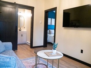 Un salon moderne avec un canapé, une petite table ronde ornée d’une plante, un téléviseur fixé au mur et une vue sur une chambre à coucher par une porte ouverte.