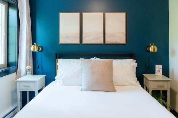 A hotel room with a bed, two bedside tables, a nightstand, a window with curtains, and three framed pictures on the wall.
