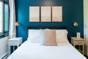 Una habitación de hotel con una cama grande, dos mesitas de noche, una lámpara de noche, una ventana con cortinas y tres cuadros en la pared.