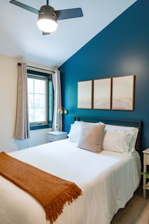 A bedroom with a bed, a ceiling fan, a window with curtains, and wall-mounted artwork.