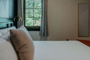 Une chambre d’hôtel avec un lit, une fenêtre avec des rideaux, une table de chevet et un miroir fixé au mur.
