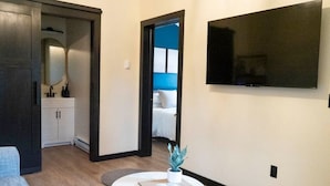 Een moderne woonkamer met een bank, een klein rond tafeltje met een boek, een potplant, een aan de muur bevestigde televisie, en een doorgang naar een slaapkamer.