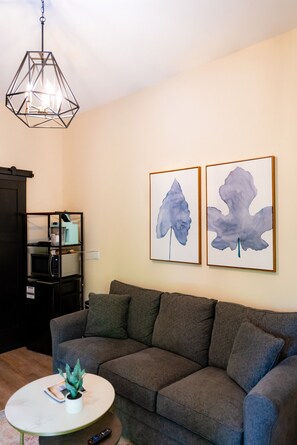 Ein Wohnzimmer mit grauem Sofa, einem weißen Couchtisch mit Pflanze, einer schwarzen Küchenzeile und zwei abstrakten Bildern an der Wand.