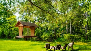 Ein Vogelhaus aus Holz hängt an einem Baum in einem üppigen, grünen Garten mit drei Stühlen und einem Tisch.