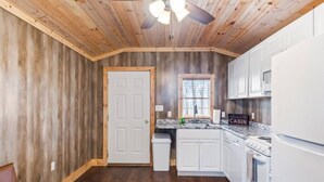 L’intérieur d’une cabane avec des murs en bois, une porte blanche, une cuisine avec des meubles blancs, un poêle et un ventilateur de plafond.