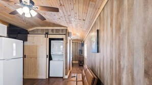 Une cuisine compacte équipée d’un réfrigérateur, d’un lave-linge et d’un ventilateur de plafond. Un banc est placé contre le mur, et une porte donne accès à une autre pièce.