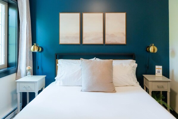 Une chambre d’hôtel avec un grand lit, deux tables de chevet, une commode, une fenêtre avec des rideaux et trois tableaux encadrés accrochés au mur.
