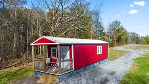 A red cabin with a porch, a small deck, and a gravel driveway. - Tiny cabin on 10 acres with pond (Albertville)