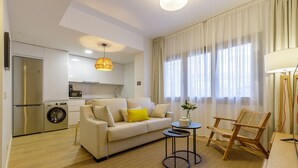 A modern living room with a sofa, a small round table, and a wooden chair. There is a kitchen area with a washing machine and a refrigerator.