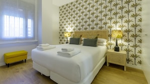 A hotel room with a large bed, a bedside table with a lamp, a small bench, and a window with sheer curtains.