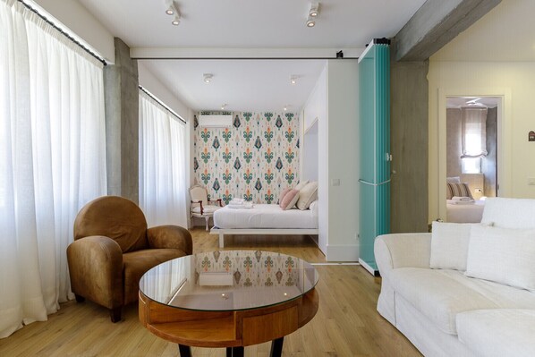 A modern hotel room with a round glass table, a brown leather chair, a white sofa, and a bed with patterned wallpaper. - Alcazaba Boutique Apartment (Málaga)
