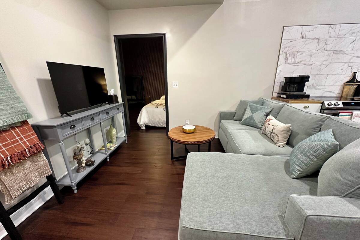 A modern living room with a grey sofa, a television on a stand, a coffee machine, and a dining area with a table and chairs.
