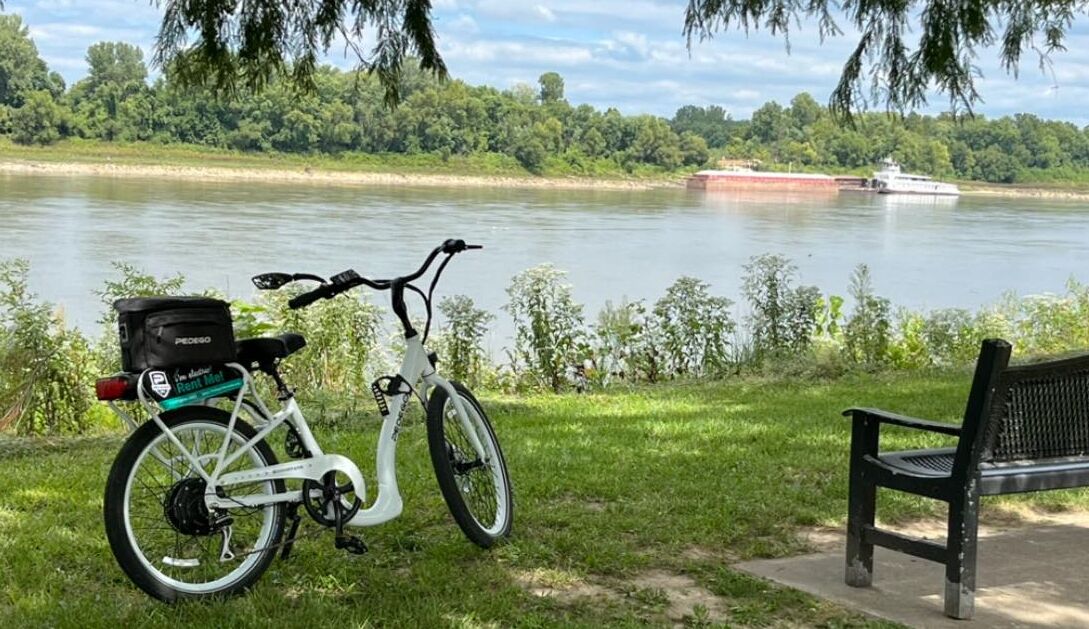 A white bicycle with a black basket and a green label, parked on grass near a river with a bench and a large boat in the background.
