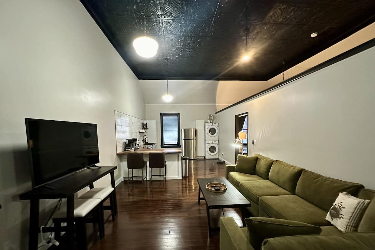 A compact living space with a flat-screen TV, a green sofa, a dining area with a table and chairs, a kitchen with a refrigerator and washing machine, and a ceiling with a light fixture.