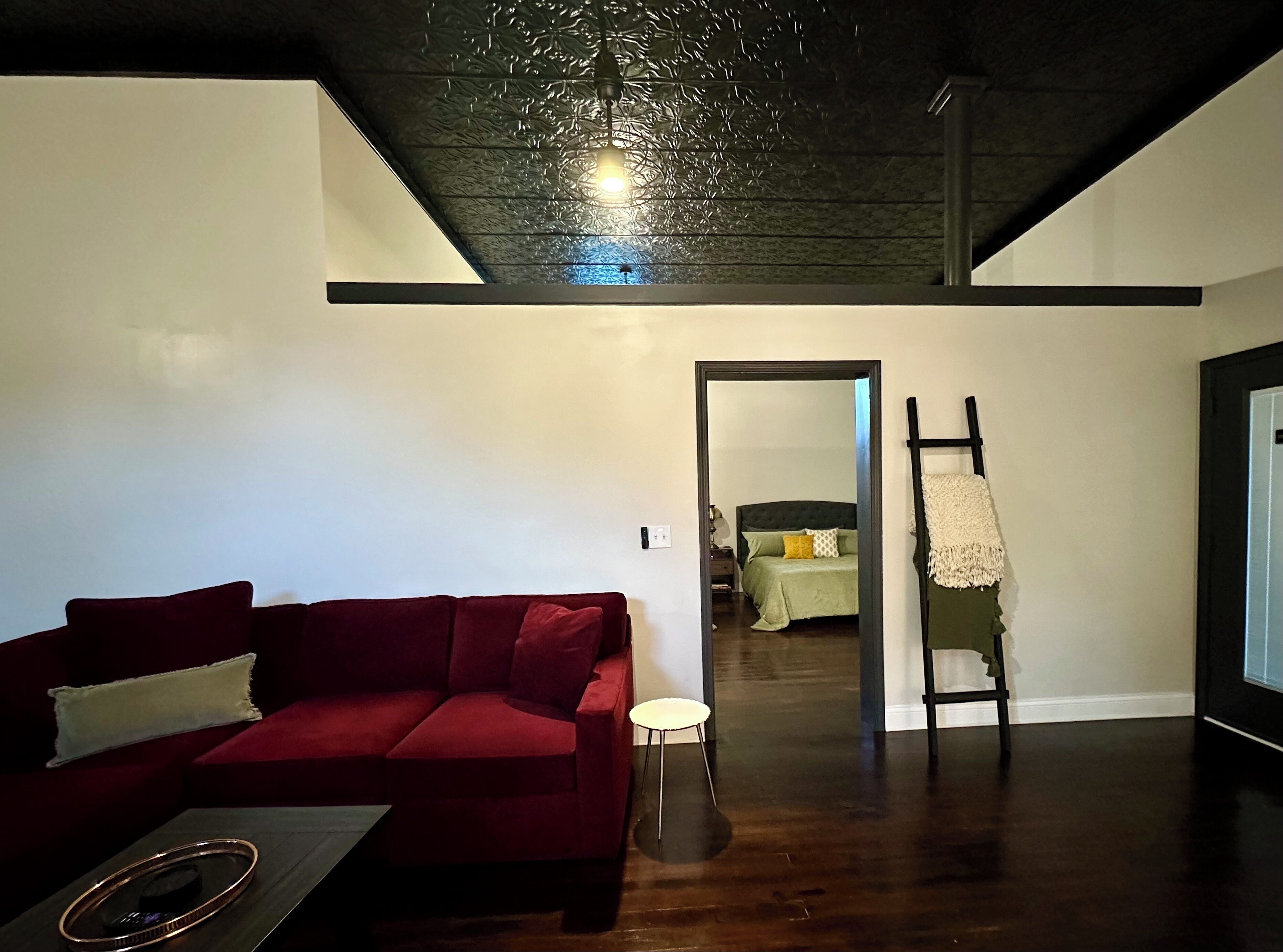 Un salon moderne avec un canapé rouge, une table basse et un plancher en bois. Une porte donne accès à une chambre à coucher où se trouvent un lit et un fauteuil suspendu.
