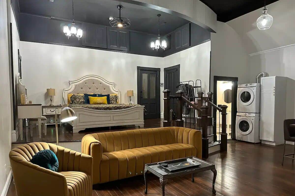 A spacious living room with a large sofa, a coffee table, and a chandelier. There is a staircase leading to another level, and a washing machine is visible in the background.