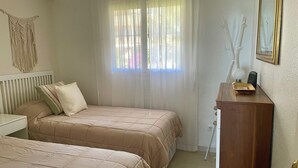 Une chambre avec deux lits, une table de chevet, une commode, une fenêtre avec des rideaux légers et une décoration en macramé accrochée au mur.