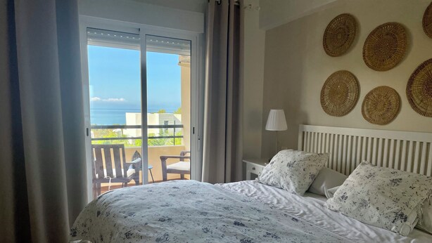 Une chambre à coucher avec un lit, une table de chevet, une lampe et une vue sur la mer, visible par la porte d’un balcon.