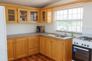 A kitchen with wooden cabinets, a stainless steel sink, a gas stove, and a window with blinds. - Mountain sea View House (St. Patrick)