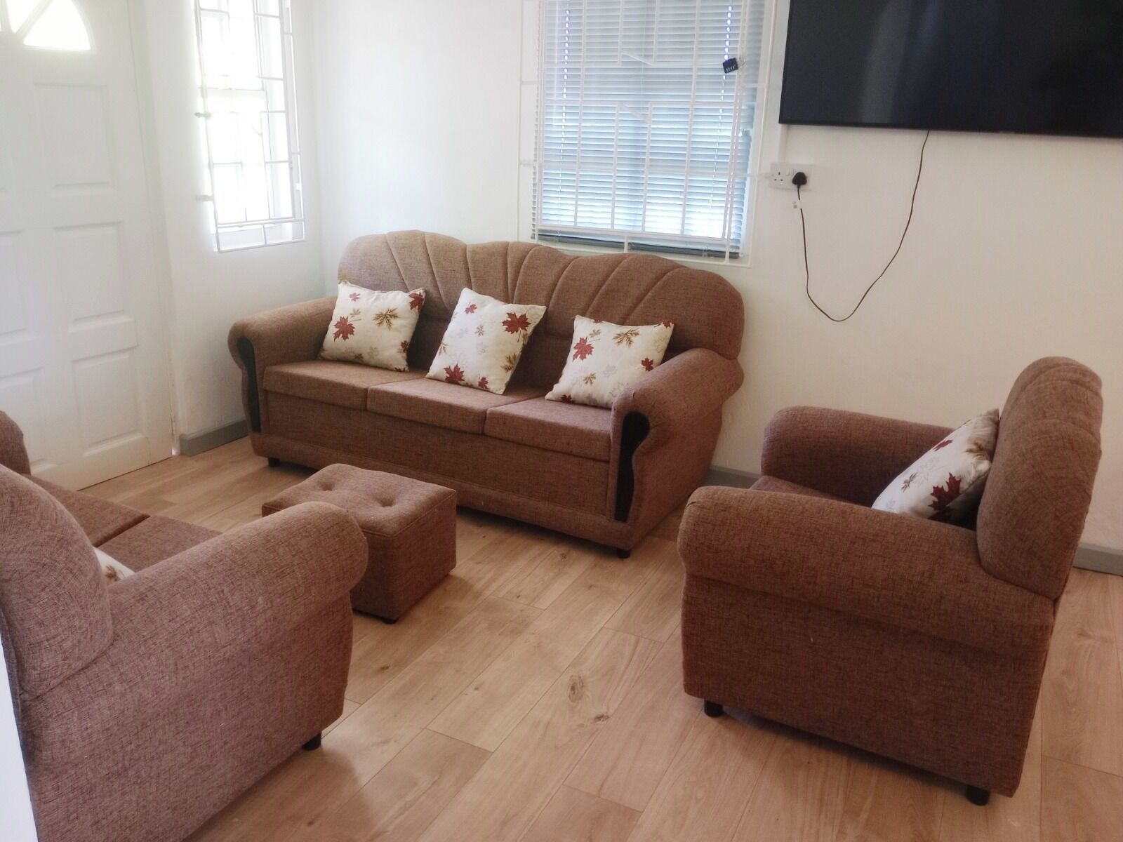 A living room with a sofa, two armchairs, and a coffee table. There is a TV mounted on the wall and a window with blinds.