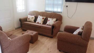A living room with a sofa, two armchairs, and a coffee table. There is a TV mounted on the wall and a window with blinds.