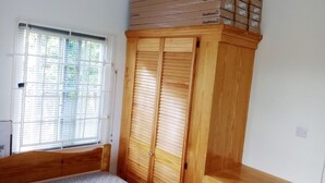Une chambre avec une armoire en bois, un lit et une fenêtre avec des stores. Des boîtes sont empilées sur le dessus de l’armoire.