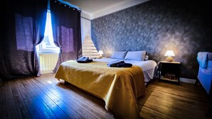 A hotel room with a wooden floor, a bed with a yellow bedspread, a nightstand with a lamp, and a view of a building outside.