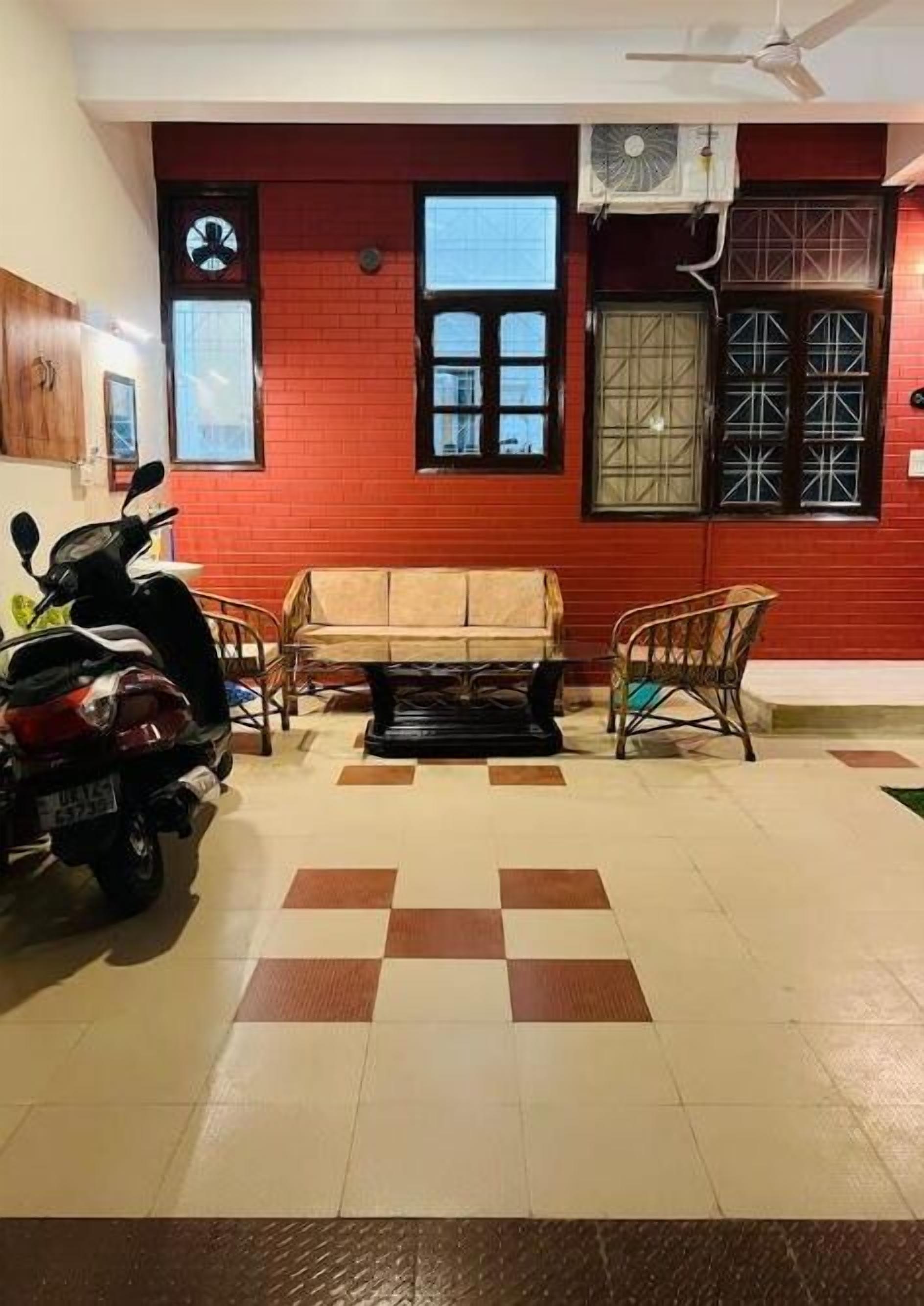 Een kamer met een rode bakstenen muur, een plafondventilator, twee ramen met metalen tralies, een bank, een stoel en een geparkeerde scooter.