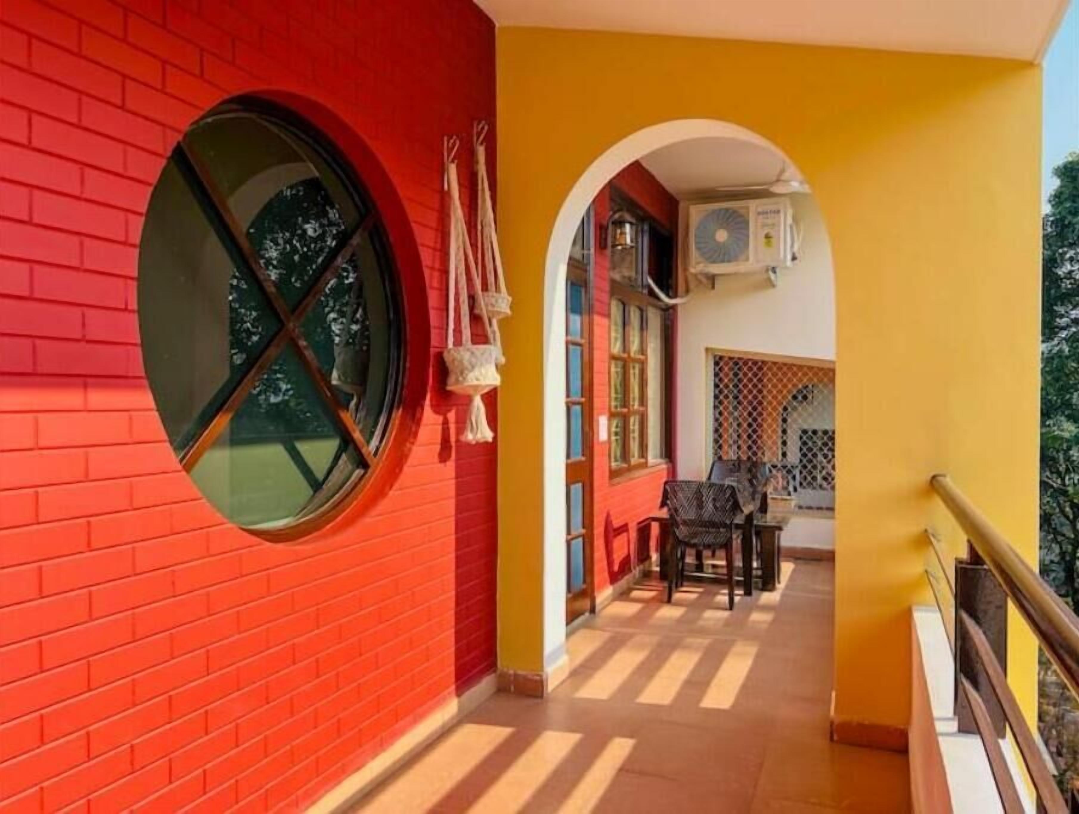 A corridor with a red brick wall, a yellow wall, and a window with a cross pattern. There is a table with chairs and an air conditioning unit mounted on the yellow wall.