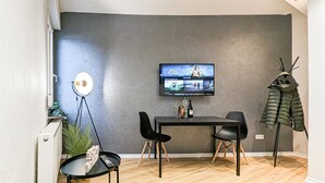 Ein modernes Wohnzimmer mit einem an der Wand montierten Flachbildfernseher, einem kleinen runden Tisch mit Fernbedienung, zwei schwarzen Stühlen und einem Kleiderständer mit einer Daunenjacke.