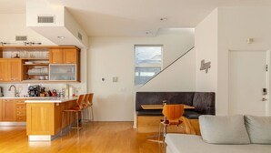 A modern kitchen with wooden cabinets, a central island, and bar stools. There is a sofa and a dining table in the living area.