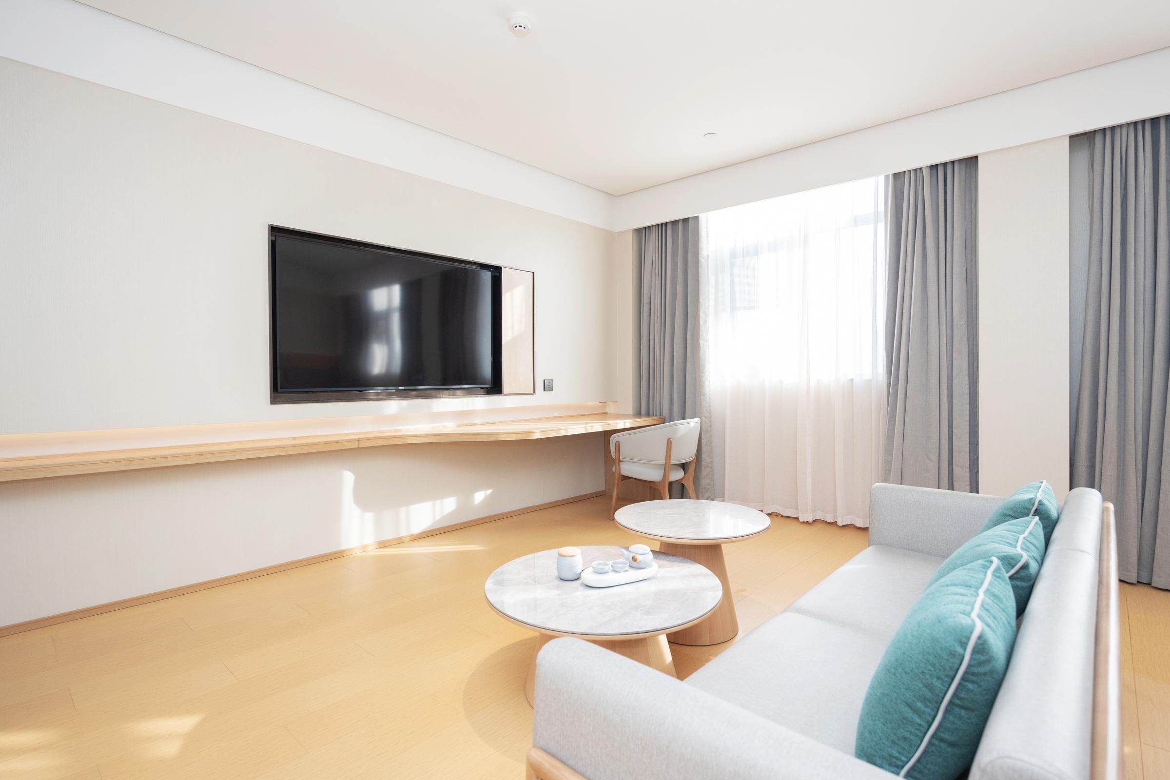 A modern hotel room with a flat-screen TV mounted on the wall, a sofa, two small round tables, and a chair.