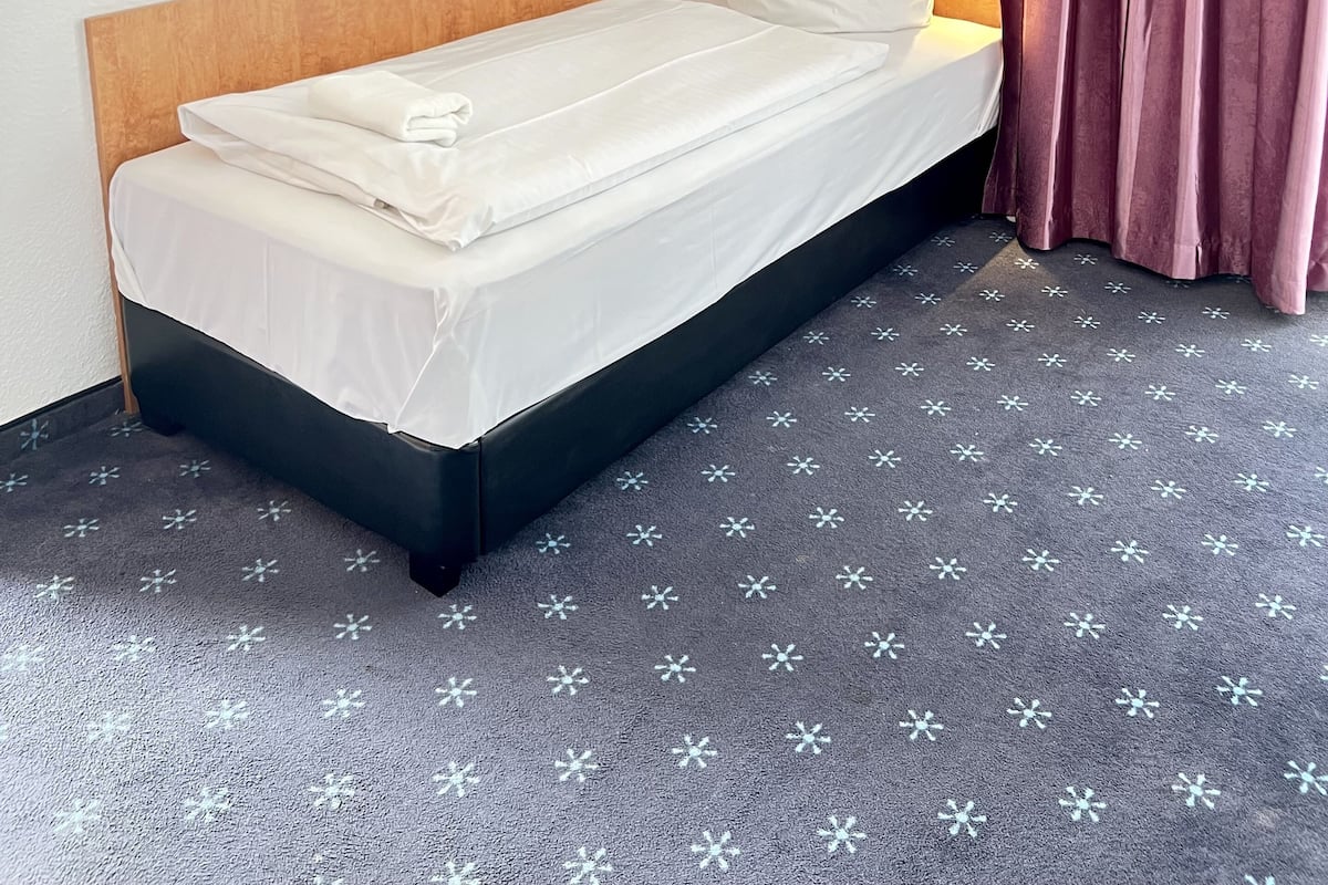 Een enkel bed met witte lakens, een houten hoofdbord, een kamerbreed tapijt met dessin, en een paars gordijn.