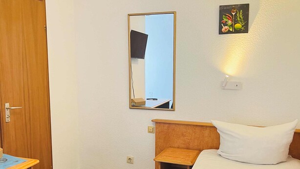 Hotellihuone, jossa on puinen ovi, pieni puinen työpöytä, valkoisilla lakanoiden peitetty sänky, yöpöytä, peili ja seinälle asennettu valaisin.
