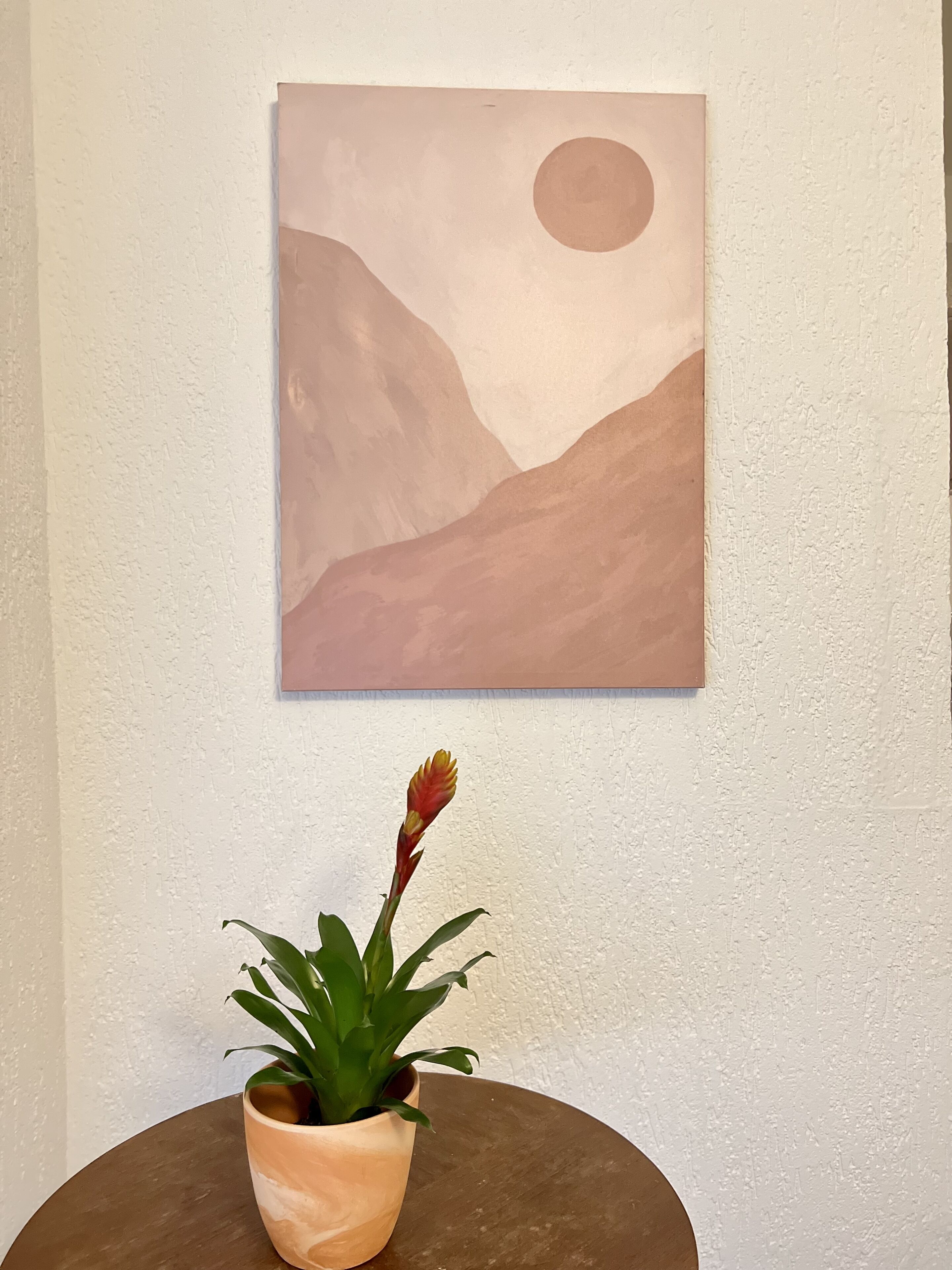 Een schilderij van een landschap met een maan en bergen, hangend aan een muur, en een bloempot op een rond bijzettafeltje eronder.