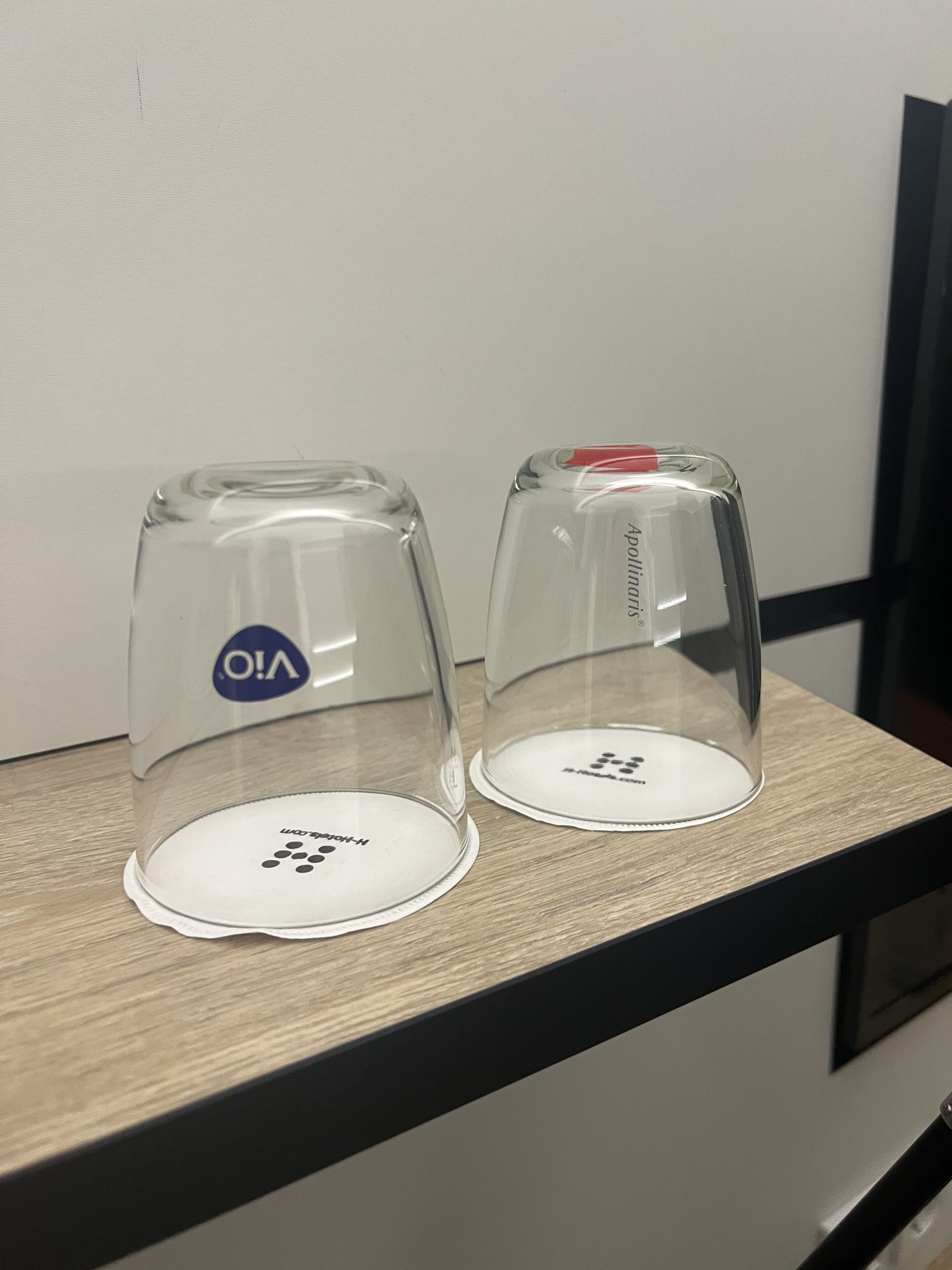 Two clear glass coffee cups with lids on a wooden table against a white wall.